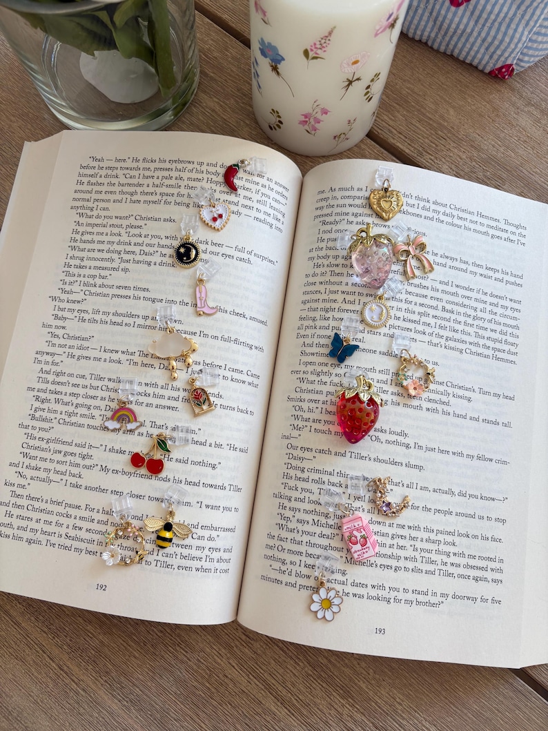 May include: A selection of charm pendants, including a strawberry and a bee, on an open book. The charms are attached to clear plastic. The book is open to pages 192 and 193. The book is on a wooden surface.