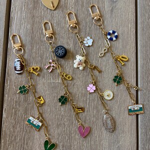 Op de afbeelding: Gouden sleutelhangers met diverse bedels, waaronder een voetbal, een teddybeer, een klavertjevier en een cassettebandje. De bedels zijn in verschillende kleuren, zoals groen, roze en wit. Er is ook een gouden hartvormige speld aanwezig.