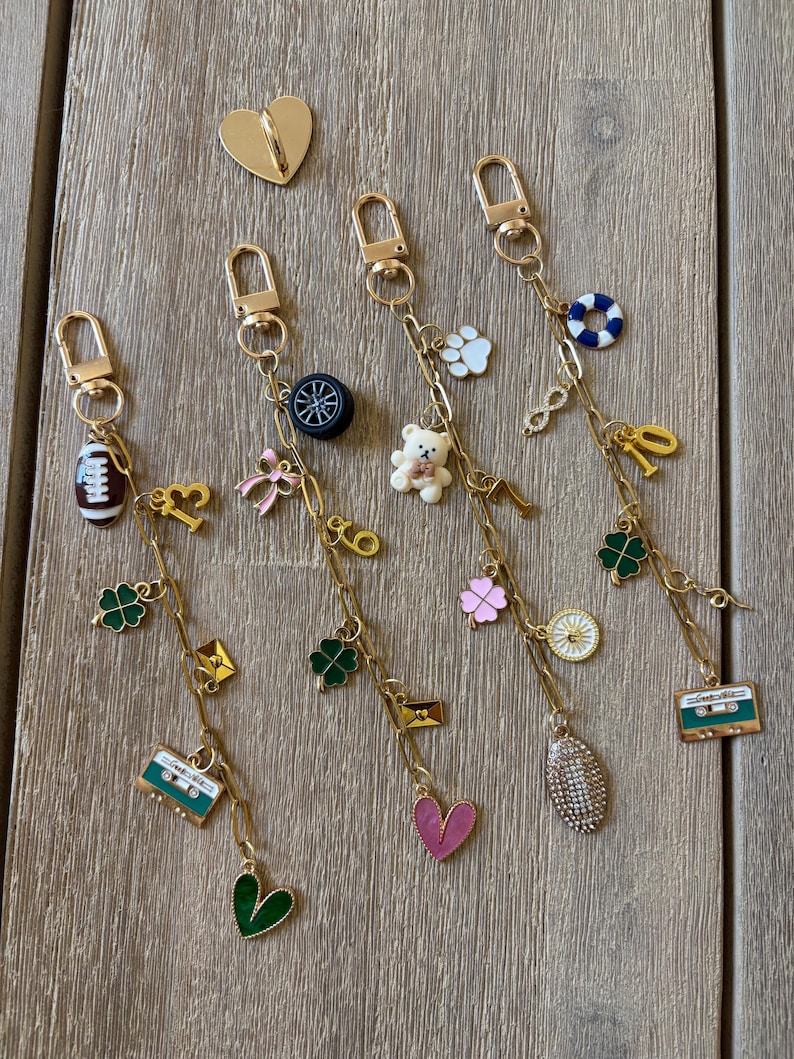 Op de afbeelding: Gouden sleutelhangers met diverse bedels, waaronder een voetbal, een teddybeer, een klavertjevier en een cassettebandje. De bedels zijn in verschillende kleuren, zoals groen, roze en wit. Er is ook een gouden hartvormige speld aanwezig.