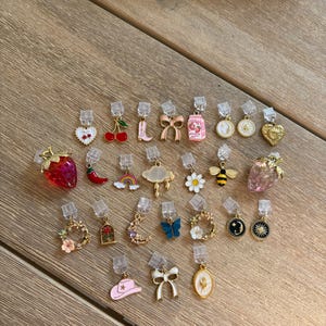 May include: A selection of clip-on earrings with diverse designs, such as strawberries, cherries, bows, and flowers. The earrings have gold-coloured metal and colourful enamel details, ideal for adding a touch of whimsy to any outfit.