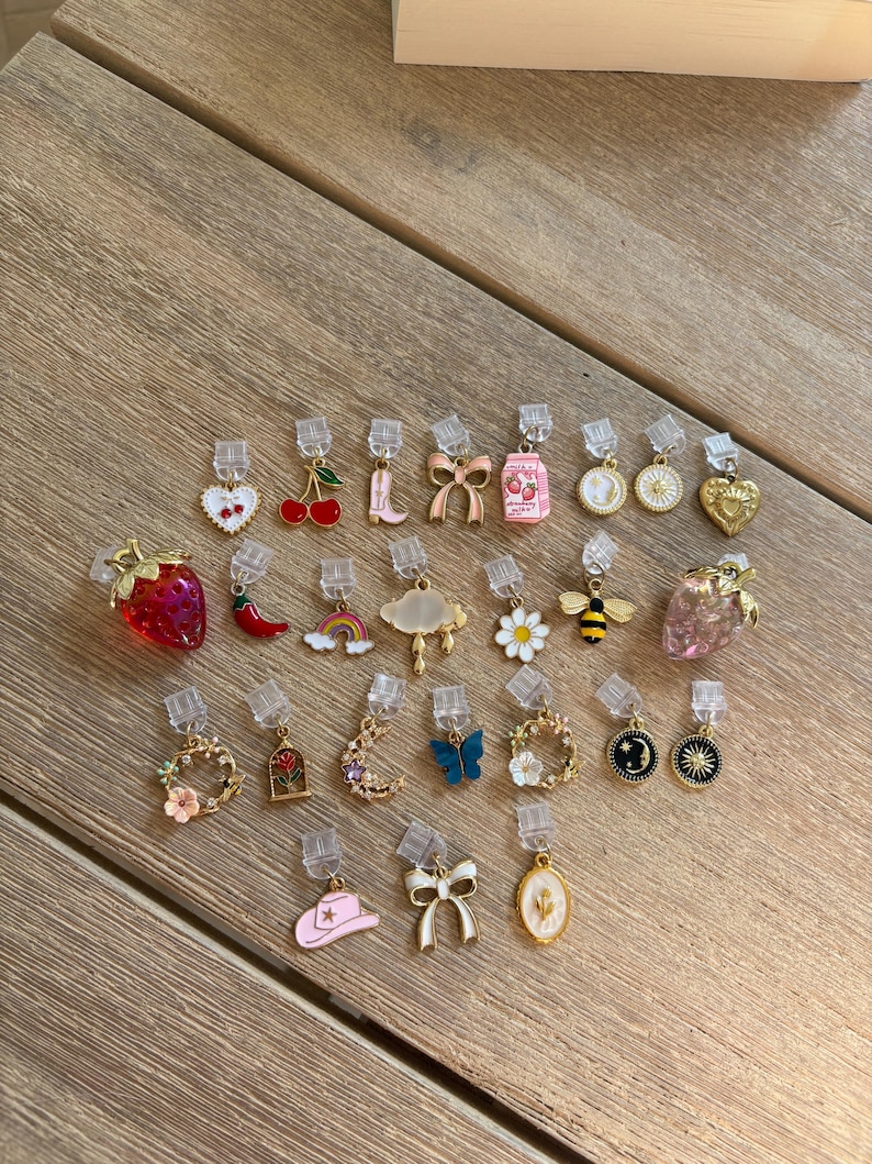 May include: A selection of clip-on earrings with diverse designs, such as strawberries, cherries, bows, and flowers. The earrings have gold-coloured metal and colourful enamel details, ideal for adding a touch of whimsy to any outfit.