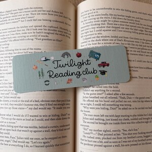 May include: A rectangular light blue bookmark featuring the text "Twilight Reading club" in black script. The design includes small illustrations of a baseball cap, bat, apple, red truck, graduation cap, trees, leaves, and other icons.