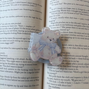 May include: A resin bear-shaped phone grip in light blue and white, featuring a blue bow and floral details. The grip sits on a light blue circle, placed on an open book.