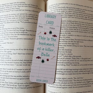 May include: A rectangular bookmark with a light grid pattern, featuring the text "LIBRARY CARD" at the top and "BOOK LOVER" at the bottom. The center displays the text "This is the bookmark of a killer, Bella" surrounded by small icons of red apples, pine trees, red trucks, and baseball bats.