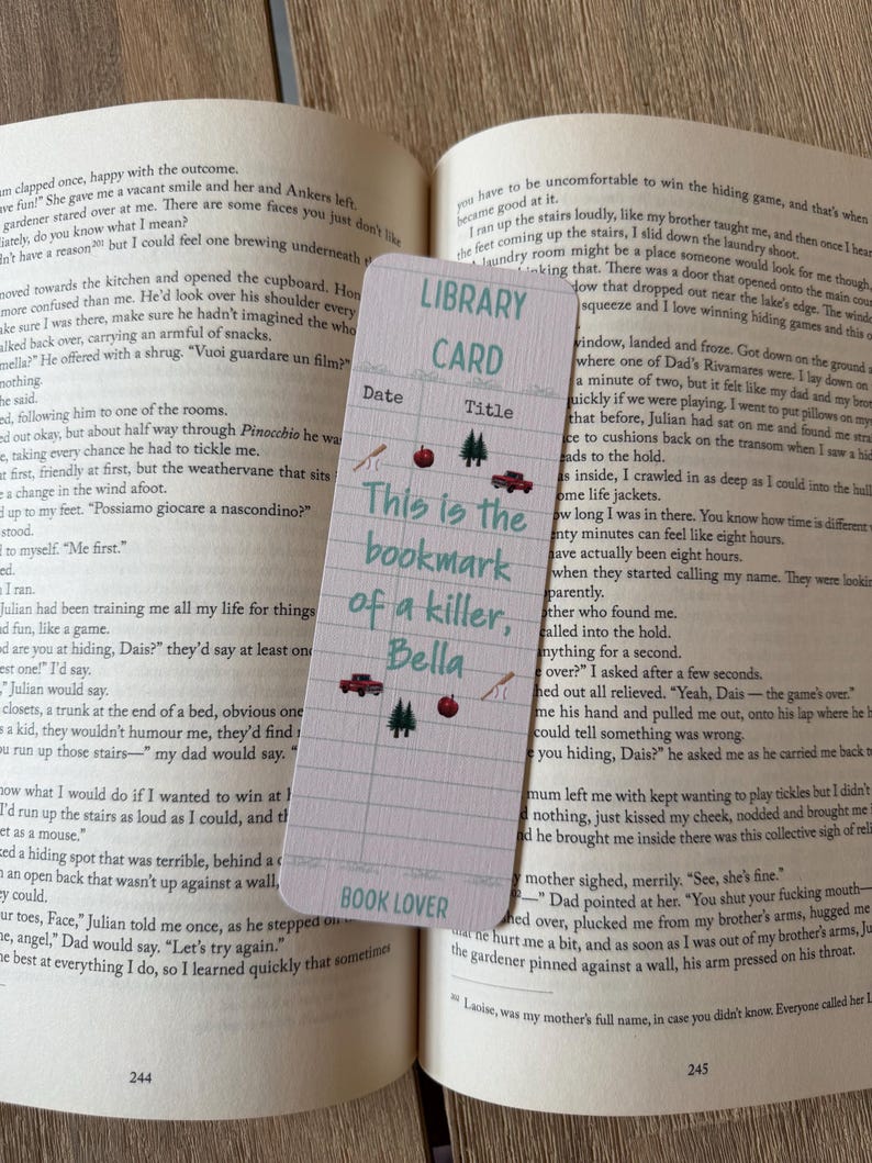 May include: A rectangular bookmark with a light grid pattern, featuring the text "LIBRARY CARD" at the top and "BOOK LOVER" at the bottom. The center displays the text "This is the bookmark of a killer, Bella" surrounded by small icons of red apples, pine trees, red trucks, and baseball bats.