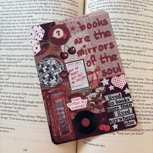 May include: A bookmark with the words "books are the mirrors of the soul." It features a red telephone box, a vinyl record, a disco ball, and cherries. The text "man written by women" is also visible on the bookmark.