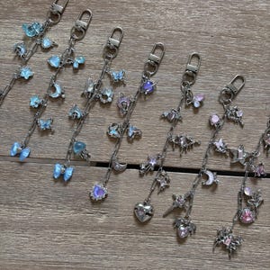 May include: A collection of silver keychains, each adorned with decorative charms. The charms feature butterfly, moon, star, and heart designs in blue, purple, and pink hues. The keychains are displayed on a wooden surface.