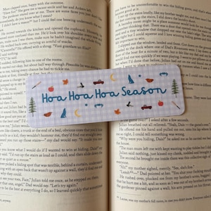 May include: A rectangular bookmark with a light blue and white gingham pattern, featuring the blue text "Hoa Hoa Hoa Season" and small icons of red trucks, pine trees, wolves, crescent moons, apples, leaves, and bats, resting on an open book.
