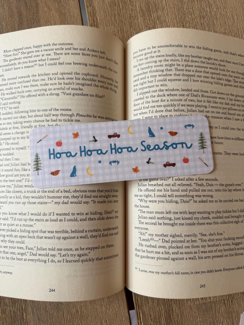 May include: A rectangular bookmark with a light blue and white gingham pattern, featuring the blue text "Hoa Hoa Hoa Season" and small icons of red trucks, pine trees, wolves, crescent moons, apples, leaves, and bats, resting on an open book.