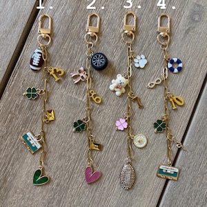 Op de afbeelding: Vier goudkleurige sleutelhangers met diverse bedels. Elke sleutelhanger heeft een karabijnhaak en een ketting met bedels zoals een voetbal, een klavertjevier, een cassettebandje, een hart, een teddybeer en een wiel. De bedels zijn in diverse kleuren.