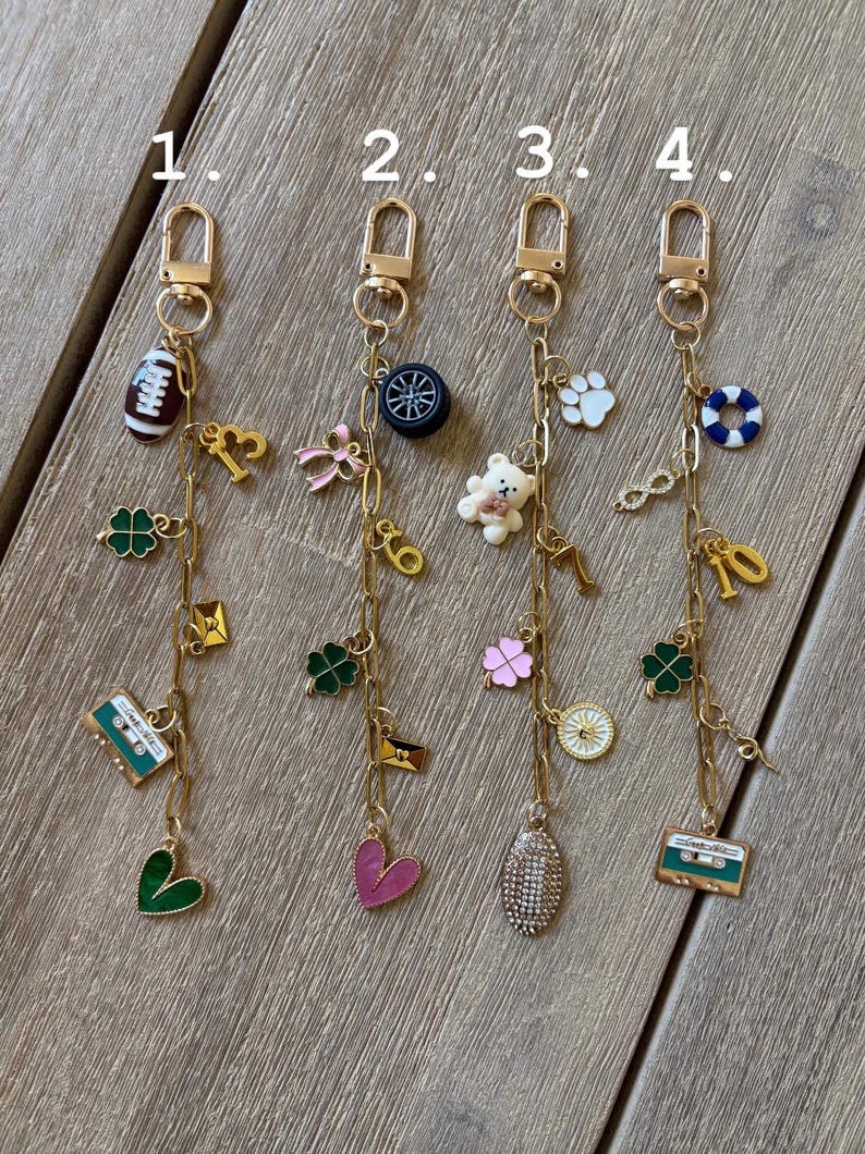 Op de afbeelding: Vier goudkleurige sleutelhangers met diverse bedels. Elke sleutelhanger heeft een karabijnhaak en een ketting met bedels zoals een voetbal, een klavertjevier, een cassettebandje, een hart, een teddybeer en een wiel. De bedels zijn in diverse kleuren.