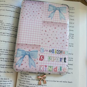 May include: Kindle case with a clear cover, featuring a patchwork design of pink gingham and floral patterns, and light blue bow illustrations. The text "EMOTIONAL SUPPORT KINDLE" is displayed in cut-out letters.