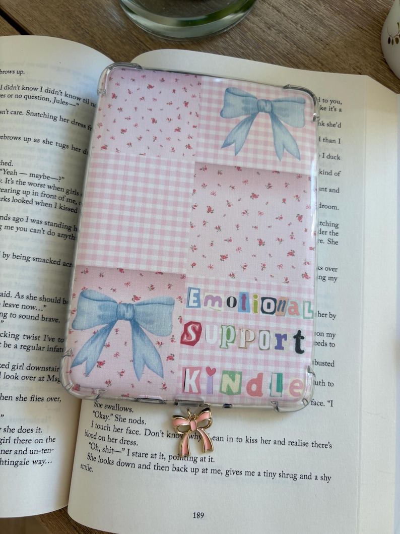 May include: Kindle case with a clear cover, featuring a patchwork design of pink gingham and floral patterns, and light blue bow illustrations. The text "EMOTIONAL SUPPORT KINDLE" is displayed in cut-out letters.