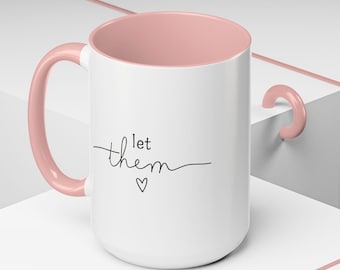 Könnte beinhalten: Weiße Keramiktasse mit rosafarbenem Innenraum und Henkel. Die Tasse hat ein schwarzes kursives Design mit dem Text "let them" und einem kleinen Herz unter dem Text.