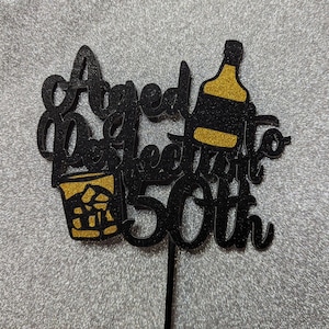 Puede incluir: Adorno para tarta negro brillante con las palabras "Aged to Perfection 50th". El diseño incluye una copa de cóctel con hielo y una botella con detalles dorados. Un palito negro está adjunto en la parte inferior.