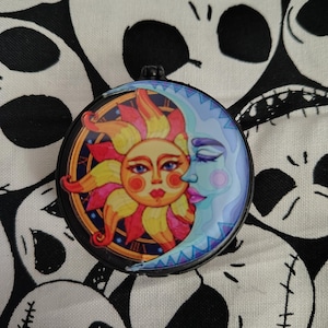 Può includere: Un specchietto compatto rotondo e nero con un design colorato di sole e luna. Il sole ha un volto con labbra rosse e raggi gialli, arancioni e rosa. La luna è azzurra con gli occhi chiusi. Lo sfondo è un motivo in bianco e nero.
