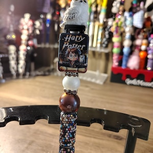 May include: A decorative pen featuring a white owl topper with a multicoloured crystal crown, a rectangular charm with the text Harry Potter and a cartoon character illustration, followed by white, brown, and multi-coloured crystal-encrusted beads.