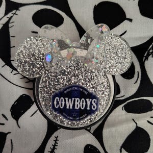 May include: A silver glitter phone grip in the shape of a mouse head with a bow. The grip has a blue and white Cowboys logo with the text "COWBOYS" and "COLD DRINKS & GOOD FRIENDS". The background is a black and white skull pattern.