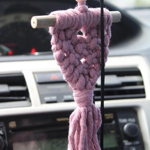 Op de afbeelding: Een handgemaakte, stoffige roze macram&eacute; auto-hanger. De hanger heeft een geweven hartvorm met een houten deuvel en een houten kraal. Een kwast hangt aan de onderkant. De hanger wordt opgehangen door een zwart koord.