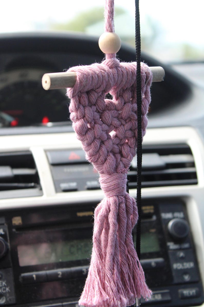 Op de afbeelding: Een handgemaakte, stoffige roze macram&eacute; auto-hanger. De hanger heeft een geweven hartvorm met een houten deuvel en een houten kraal. Een kwast hangt aan de onderkant. De hanger wordt opgehangen door een zwart koord.
