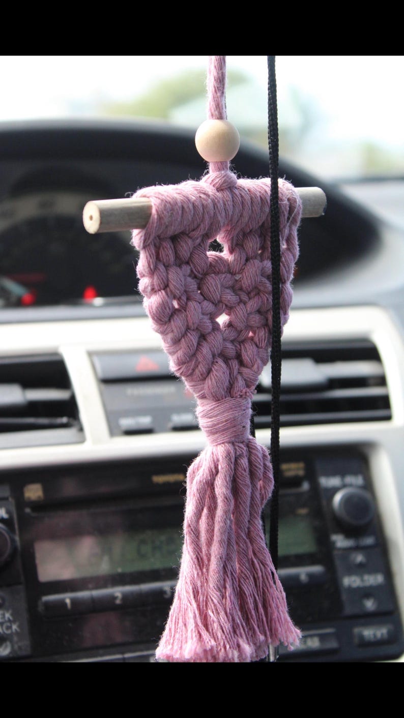 Op de afbeelding: Een handgemaakte macram&eacute; auto-hanger in een stoffige roze kleur. De hanger heeft een geweven hartontwerp, een houten deuvel en een kwast. Bovenop zit een houten kraal. Hij hangt aan een zwart koord.