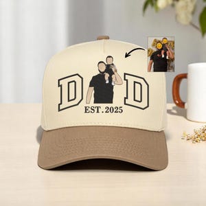 May include: A beige and brown baseball cap with the word "DAD" in black block letters and the text "EST. 2025". The cap features a graphic of a father and child. A small image of the father and child is in the upper right corner.