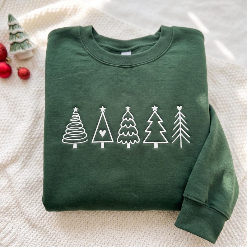 May include: A dark forest green crewneck sweatshirt featuring a white embroidered graphic of five distinct stylized Christmas trees arranged in a horizontal row across the chest, each with a unique line design and a small star or heart topper.