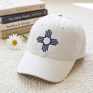 Puede incluir: Una gorra de béisbol blanca con un símbolo solar Zia bordado en azul oscuro en la parte delantera. La gorra tiene una corona estructurada, una visera curva y ojales cosidos para la ventilación, presentando un diseño limpio y sencillo.