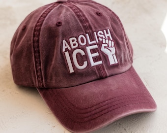 May include: A burgundy baseball cap with the text "ABOLISH ICE" embroidered in white, along with a white fist graphic. The cap has a curved brim and a distressed, vintage look.