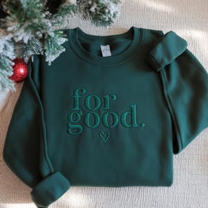 May include: A dark green sweatshirt with the embroidered phrase "for good." A small heart is embroidered below the word "good." The sweatshirt has a crew neck and long sleeves. A Christmas tree branch and ornament are in the background.