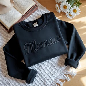 May include: A black sweatshirt with the embroidered word "Memaw" in a cursive font. The sweatshirt is folded on a white textured surface, with a book and flowers in the background.