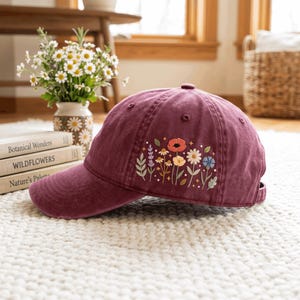 May include: A deep maroon baseball cap featuring an embroidered design of colorful wildflowers on the side panel. The embroidery includes small red, yellow, white, and blue flowers with green stems and leaves, accented by tiny star-shaped details.