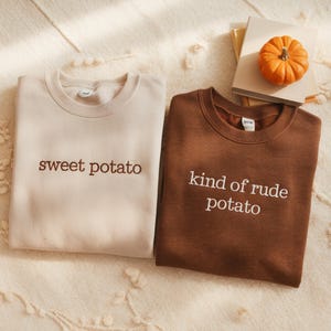 May include: Two crew neck sweatshirts, one cream and one brown, with embroidered text. The cream sweatshirt says "sweet potato" in brown. The brown sweatshirt says "kind of rude potato" in white. A small pumpkin and books are in the background.