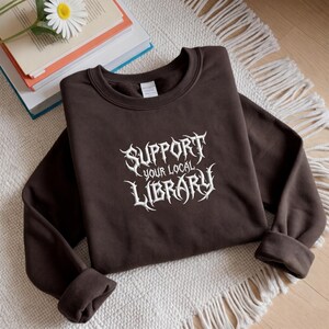 May include: A dark brown crewneck sweatshirt featuring white embroidered text on the chest that reads SUPPORT YOUR LOCAL LIBRARY in a stylized, sharp-edged font. The garment has long sleeves with ribbed cuffs and a ribbed neckline.