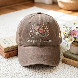 May include: A brown baseball cap with a floral design and the text "be a good human." The cap features a curved brim and a button on top. The floral design includes pink, white, and yellow flowers.