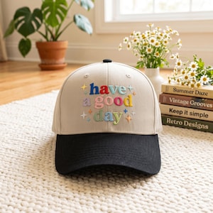 May include: A beige baseball cap with a black brim featuring the embroidered text "have a good day" in multi-colored letters. The design includes small embroidered star shapes in various colors surrounding the text on the front panel.