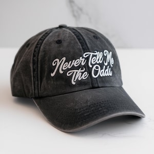 May include: A black, distressed baseball cap with the embroidered phrase "Never Tell Me The Odds" in white script. The cap has a curved brim and a fabric strap closure. The hat is made of a soft, worn material.