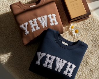 May include: A brown crewneck sweatshirt and a navy blue crewneck sweatshirt, both featuring the white embroidered text YHWH in a collegiate font. The sweatshirts are displayed alongside a tan book and two white daisy flowers with yellow centers.