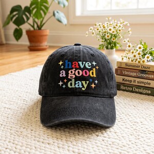 May include: A black baseball cap with an embroidered design on the front that reads "have a good day" in colorful, multi-toned letters. The text is surrounded by small, embroidered star shapes in various colors, including yellow, blue, and orange.