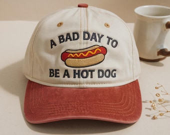 May include: A cream-colored baseball cap with a red brim and a top button. The front features embroidered text that reads A BAD DAY TO BE A HOT DOG, centered above a graphic of a hot dog with a red sausage, yellow mustard, and a tan bun.