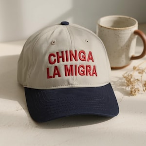 May include: A baseball cap with a light gray crown and a navy blue brim. The cap features the embroidered text "CHINGA LA MIGRA" in red. A ceramic mug with a brown handle is in the background.