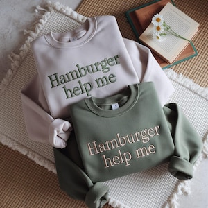 May include: Two crewneck sweatshirts, one beige and one olive green, are displayed. The beige sweatshirt has green embroidered text that reads "Hamburger help me." The olive green sweatshirt has pink embroidered text that reads the same. The sweatshirts are folded.