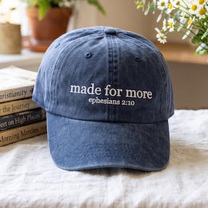 Könnte beinhalten: Eine marineblaue Baseballkappe mit verwaschener Optik, die auf der Vorderseite den in Weiß gestickten Text "made for more" gefolgt von "ephesians 2:10" zeigt. Die Kappe hat eine strukturierte Krone, einen gebogenen Schirm und einen Knopf oben.