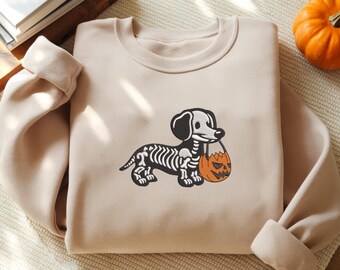 May include: A beige crewneck sweatshirt featuring an embroidered graphic of a dachshund dog with a white skeleton pattern on its body. The dog is depicted holding an orange jack-o'-lantern pumpkin bucket in its mouth, designed for Halloween.