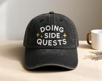 May include: A black baseball cap with the embroidered text "DOING SIDE QUESTS" in white. The text is accented with yellow starburst graphics. The cap has a curved brim and a button on top.