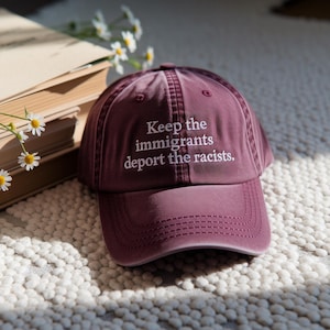 May include: A maroon baseball cap with the embroidered text "Keep the immigrants deport the racists." The hat is made of a soft material and has a curved brim. The hat is sitting on a textured surface.