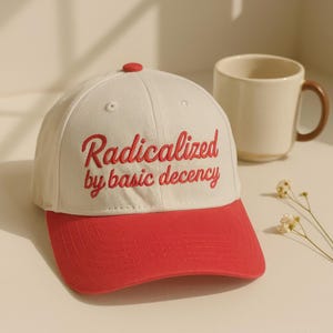 May include: A cream-colored baseball cap with a red brim and a red button on top. The front of the cap features the embroidered red text "Radicalized by basic decency" in a cursive script font. The cap has stitched eyelets on the crown.