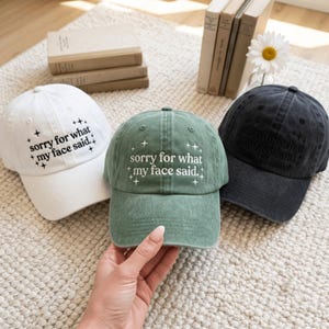 May include: Three baseball caps in white, sage green, and charcoal gray. Each cap features the embroidered text 'sorry for what my face said.' surrounded by small star icons. The white and green caps have black and white text respectively.