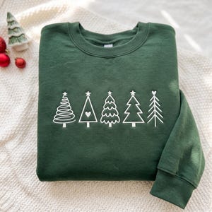 May include: A dark forest green crewneck sweatshirt featuring a white embroidered graphic of five distinct stylized Christmas trees arranged in a horizontal row across the chest, each with a unique line design and a small star or heart topper.