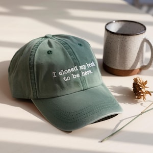 May include: A sage green baseball cap with the embroidered text "I closed my book to be here." A speckled ceramic mug with a cork base sits nearby. The cap is made of a soft fabric.